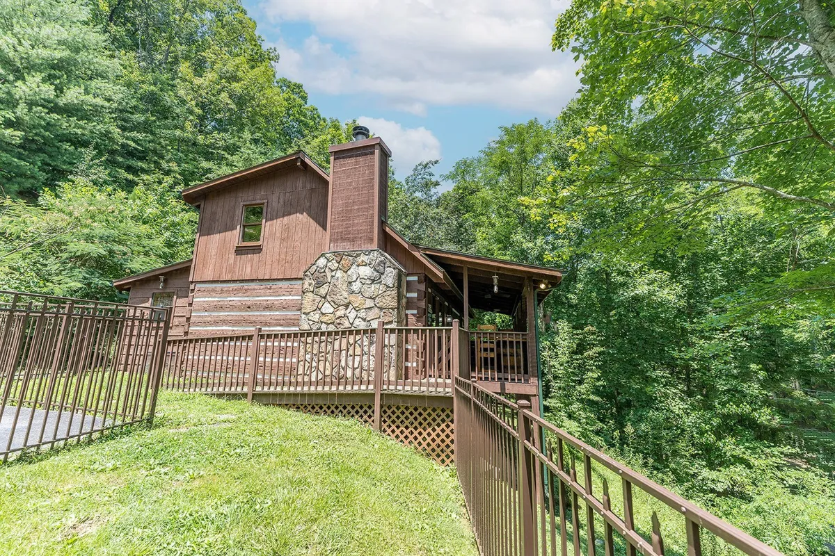 This image features the exterior of the cabin with a spacious deck area, wooden structure, stone chimney, and lush greenery surrounding the property. The deck is visible as a welcoming space for outdoor gatherings and relaxation, highlighting the property's connection to nature.