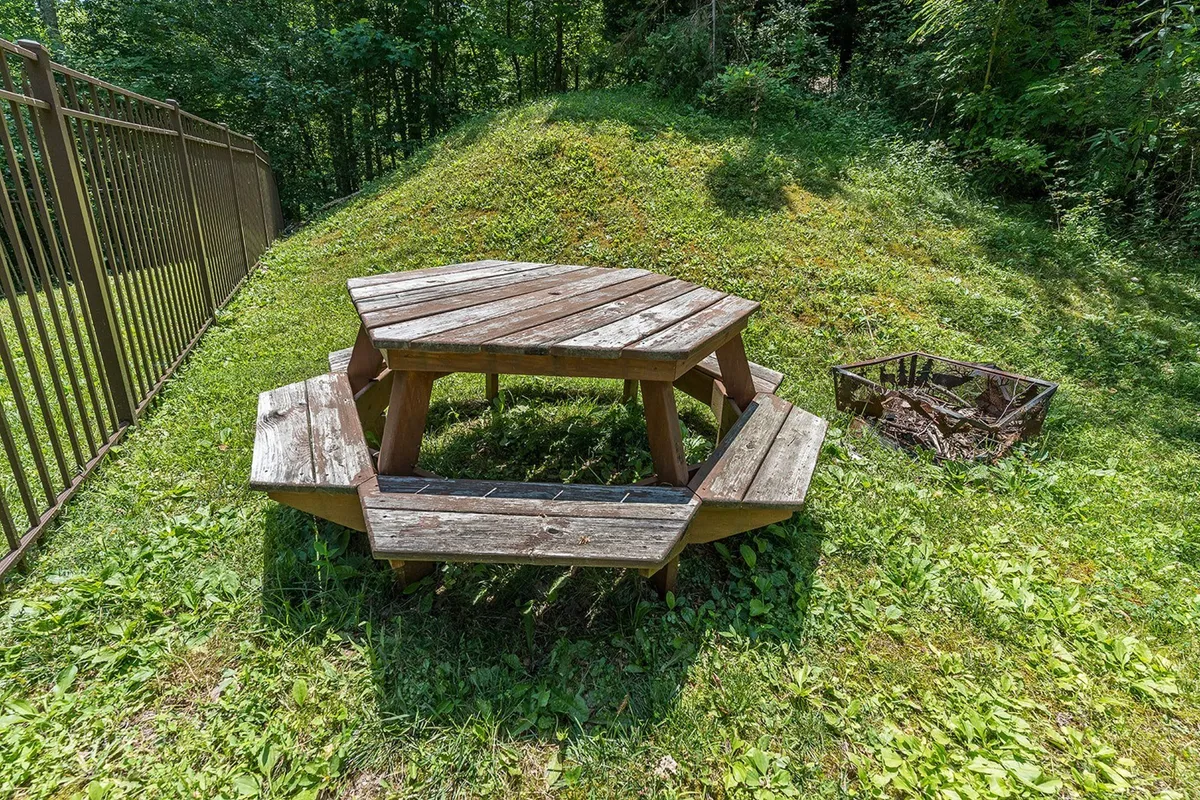 A peaceful outdoor area featuring a wooden picnic table, a cozy fire pit, and lush greenery all around. The setting is ideal for family picnics, marshmallow roasting, or relaxed stargazing around the firepit.