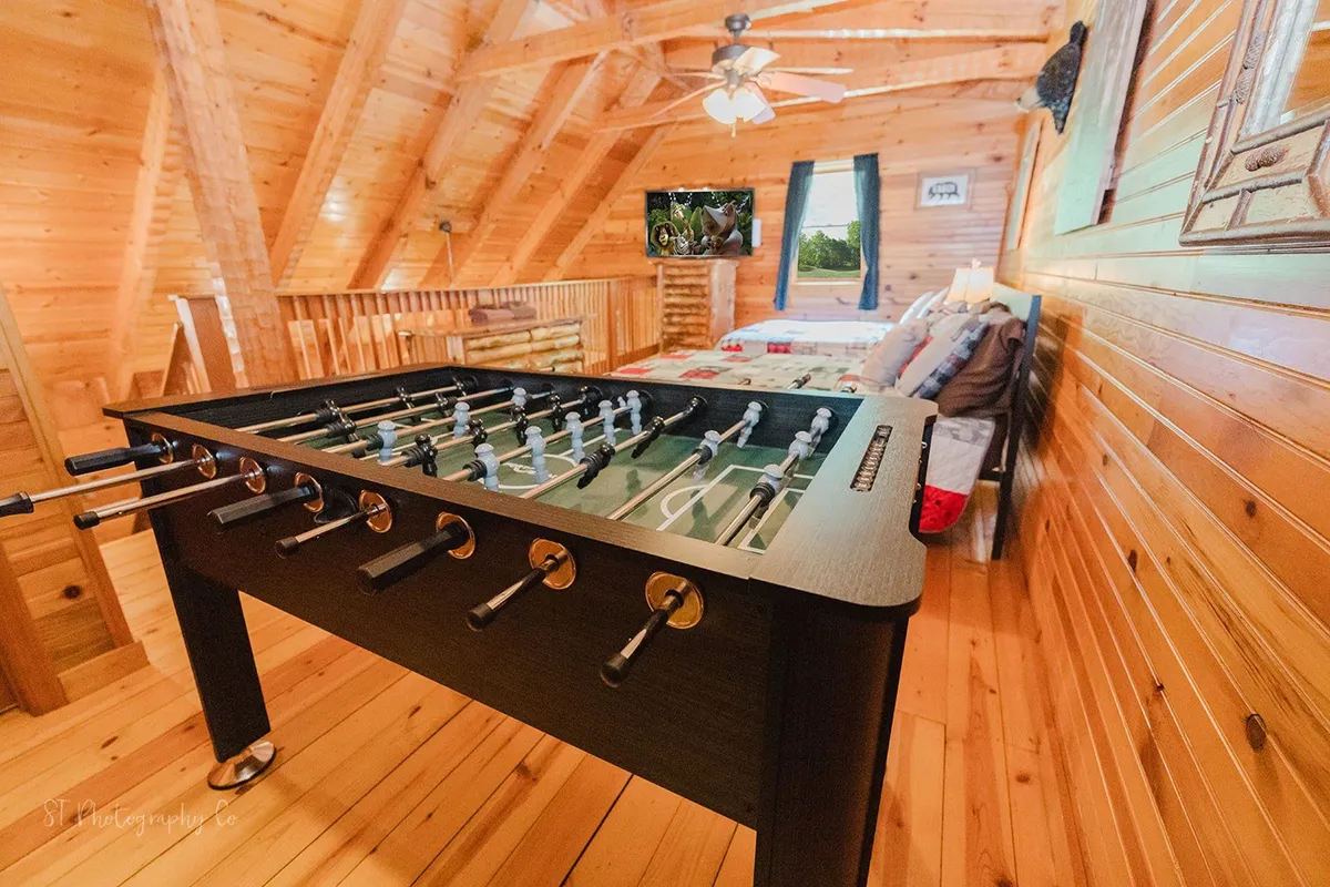 A playful loft space with a foosball table positioned near a cozy bed, complemented by a television and rustic wooden decor. The setup embodies a fun, communal sleeping area designed for group activities and friendly competition.