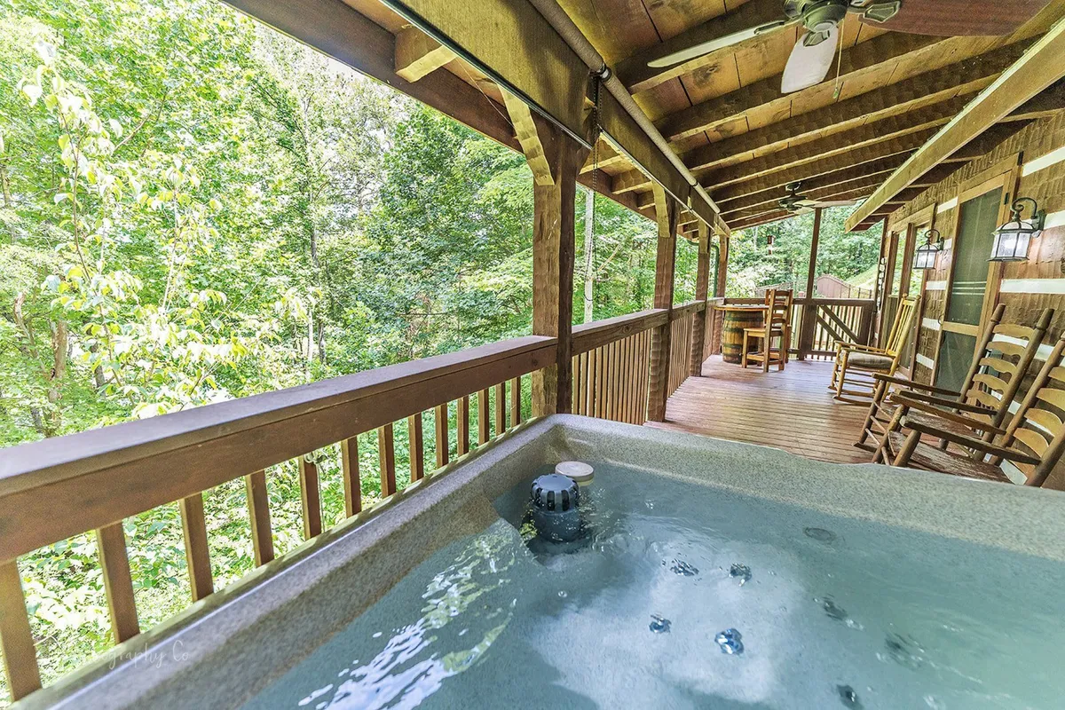 A bubbling hot tub sits on a spacious wooden deck, surrounded by lush greenery and flanked by rustic rocking chairs. The tranquil setting invites guests to relax and socialize while enjoying the sights and sounds of nature.