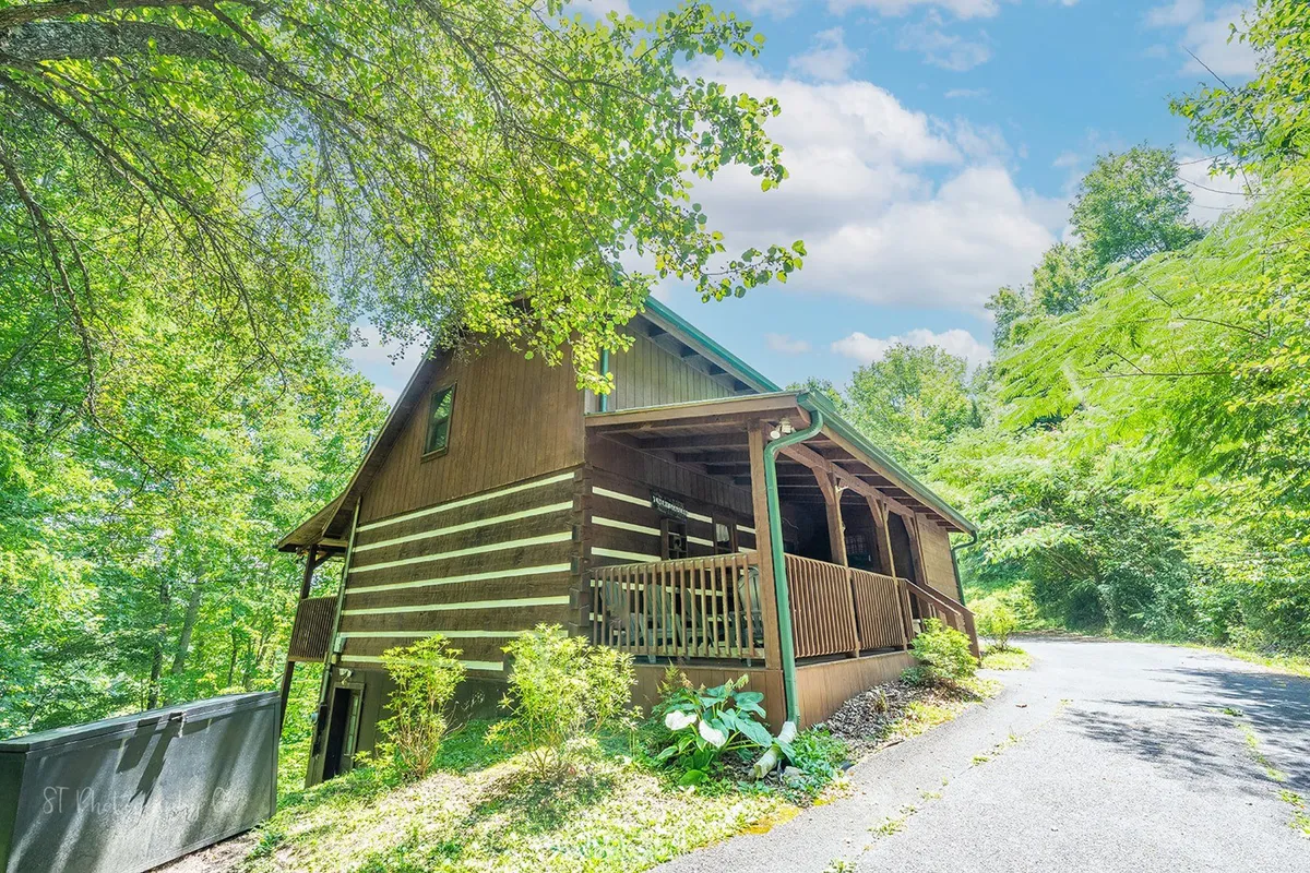 A welcoming wooden cabin sits nestled among lush greenery, with a spacious front porch ideal for relaxing. The cabin's rustic charm is highlighted by warm wood tones and inviting outdoor spaces that connect the home to its natural surroundings.