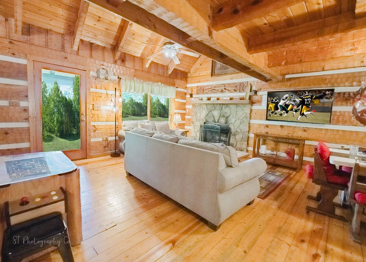 A spacious open-plan living area with a comfortable sofa set near a striking stone fireplace, a large flat-screen TV, and a dining table with chairs bathed in natural light from expansive windows. The rustic wood details and welcoming atmosphere make it an ideal gathering space.