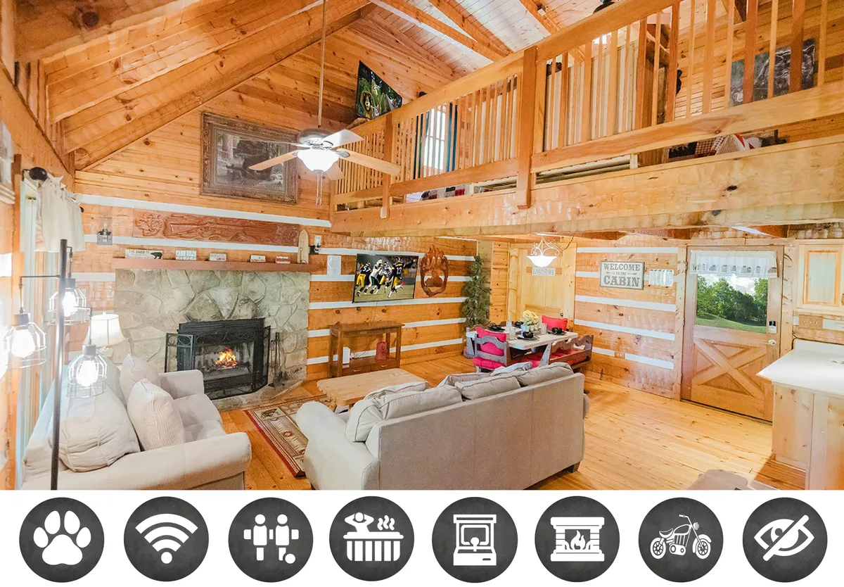 The image showcases a warm and inviting cabin interior featuring a grand stone fireplace flanked by plush, comfortable seating. Rustic wooden beams frame the space, with a cozy loft area visible above. The scene radiates comfort and togetherness, perfect for relaxing by the fire or gathering with family and friends.