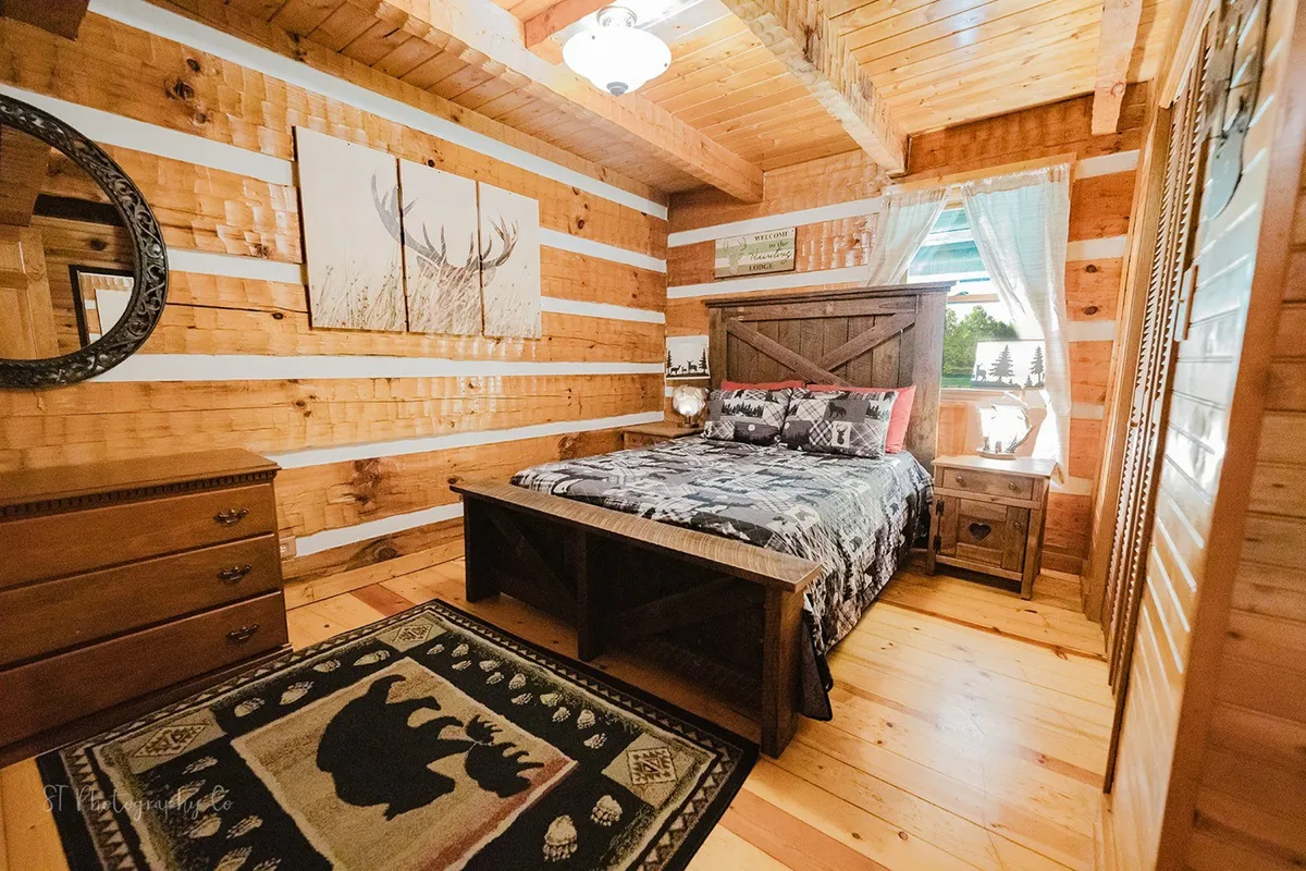 A warmly lit master bedroom featuring a wooden bed frame with plush bedding, a decorative rug, a dresser, and large windows adorned with sheer curtains that filter natural light. The room exudes an inviting, peaceful ambiance, ideal for relaxation after a day of activities.