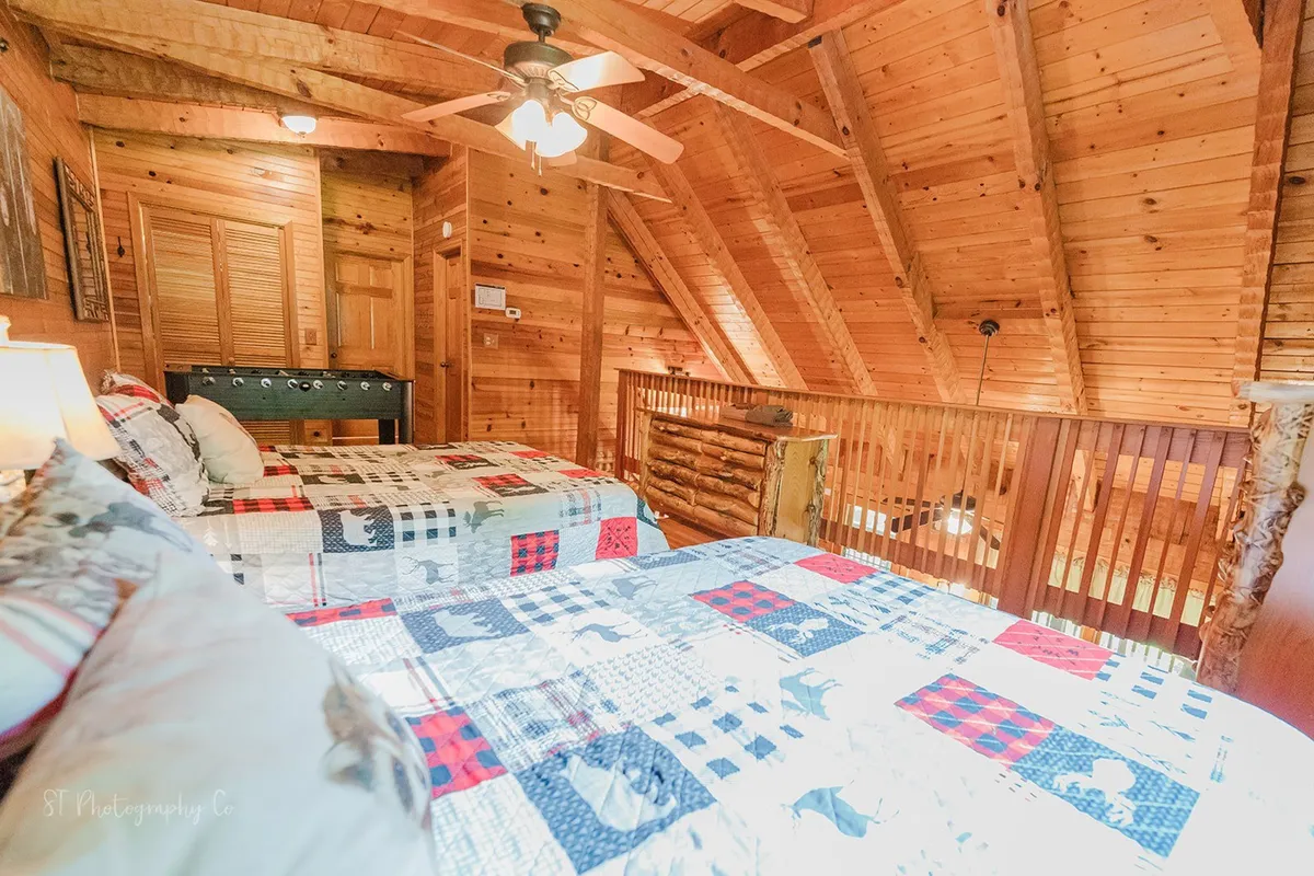 A charming loft area with two beds featuring patterned quilts, exposed wooden beams, a ceiling fan, and railings overlooking the space below. The ambiance is warm and nostalgic, inviting group gatherings and shared stories.