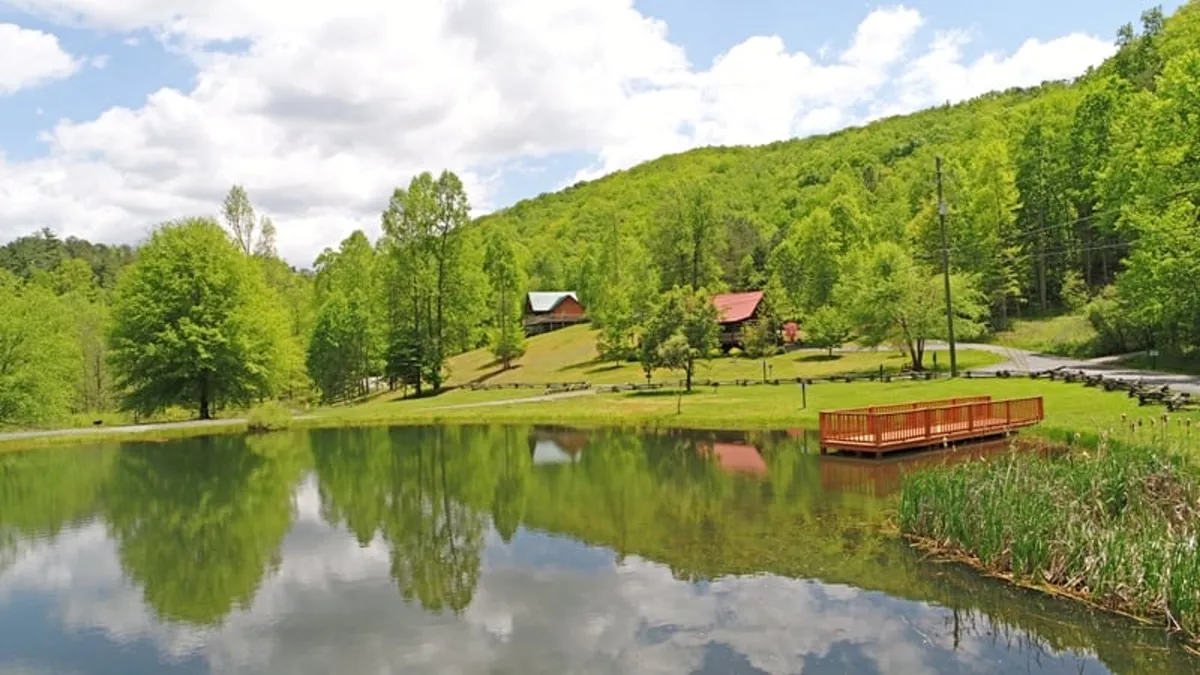 A serene pond edged by a wooden deck, lush greenery, and distant cabins in view. The scene offers a tranquil setting for group picnics, nature walks, or simply relaxing by the water.