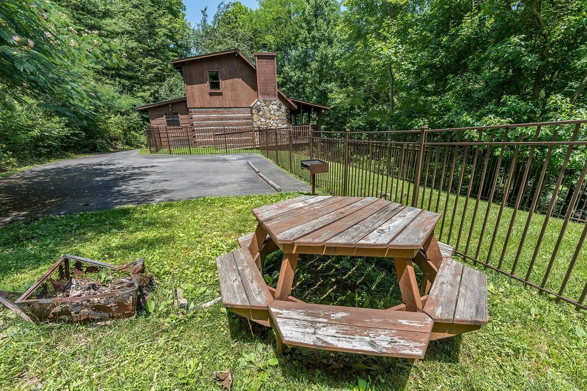 A wooden picnic table sits within a fenced yard, with the house in the background. The space is set up for outdoor dining, family picnics, and gatherings surrounded by nature.